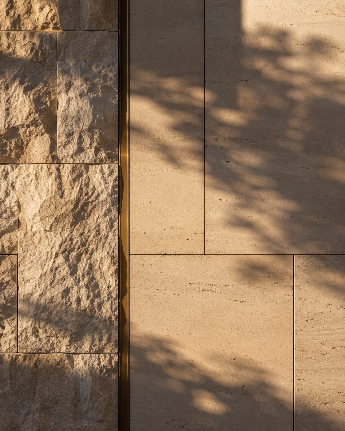 A hand-laid limestone and travertine wall in low golden light, a slim brass reveal line and the soft shadow of a tree across the stone.